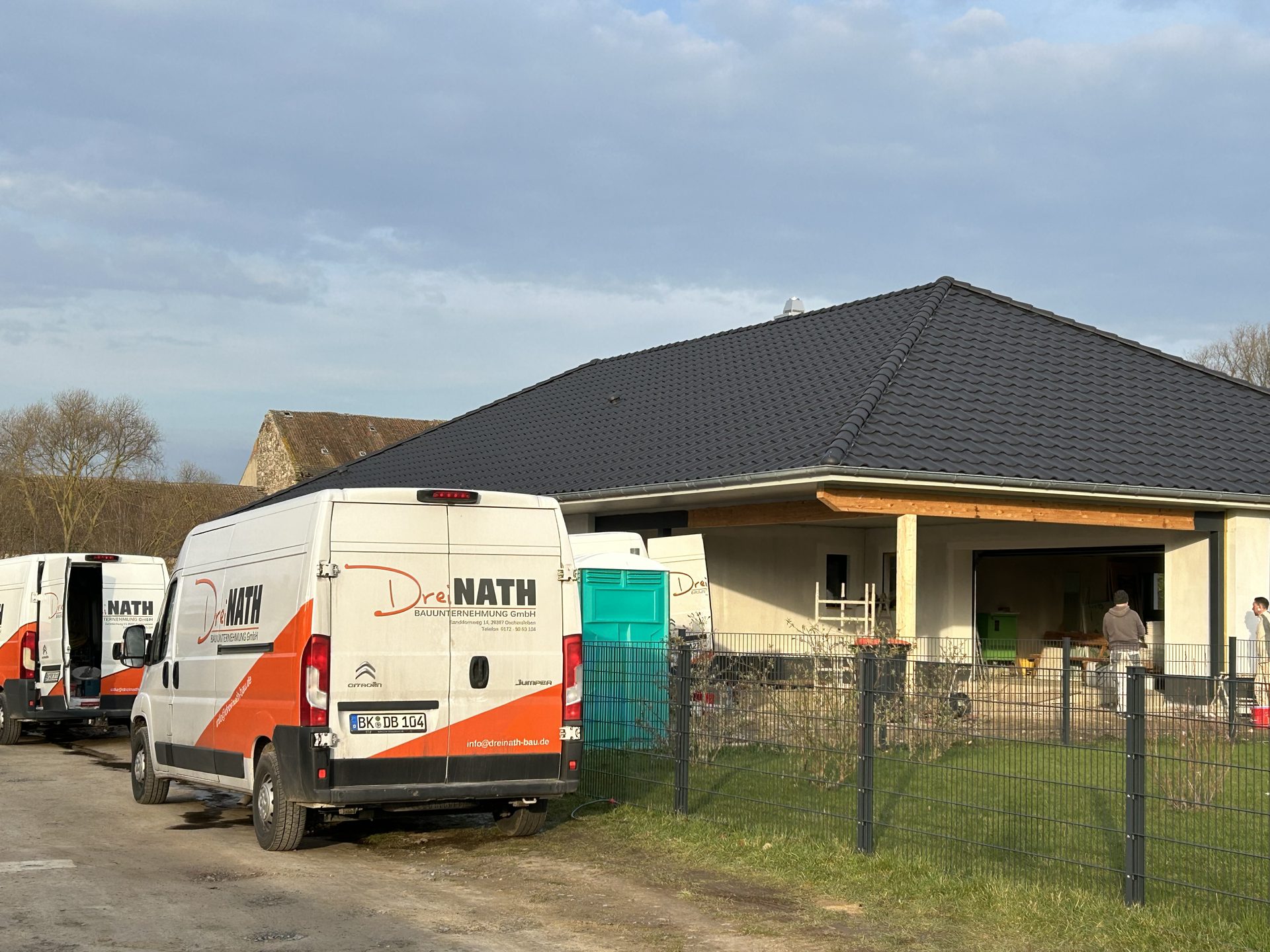 Derselbe Neubau als fertiger Bungalow mit gedecktem Dach und Firmenfahrzeugen davor